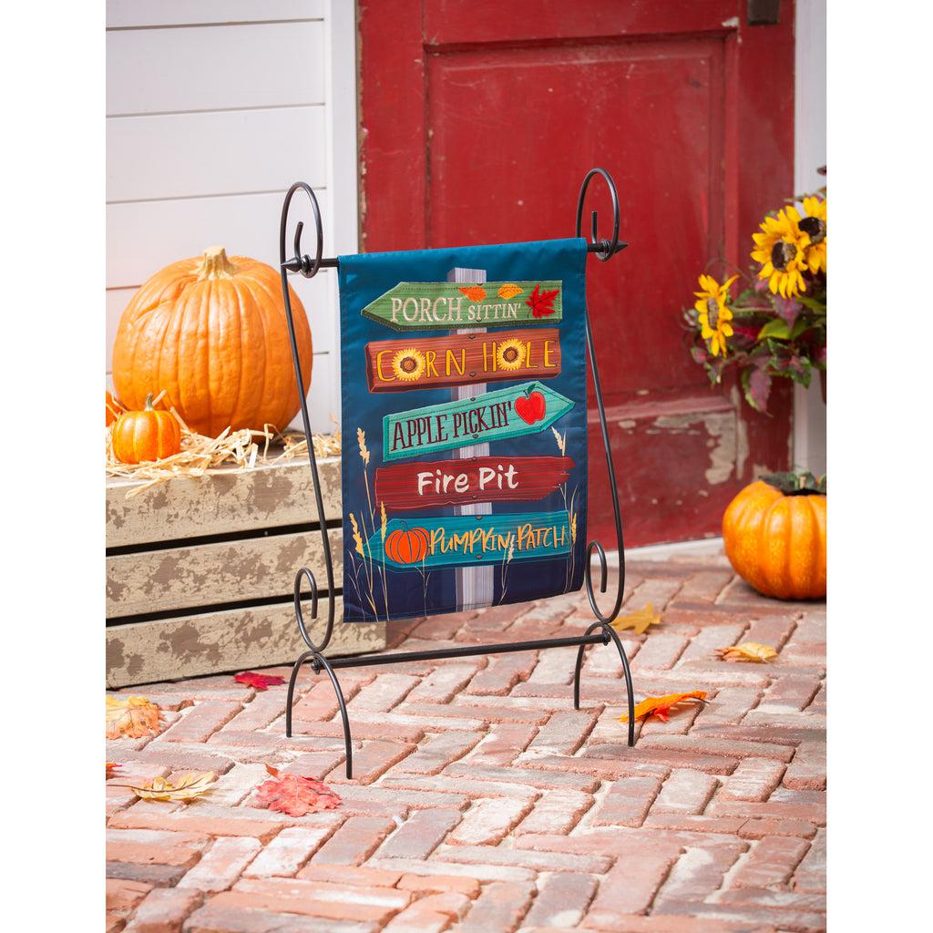 The Fall Signs garden flag features wooden arrows pointing in two different directions sending you to "Porch Sittin', Corn Hole, Apple Pickin', Fire Pit, and Pumpkin Patch". 