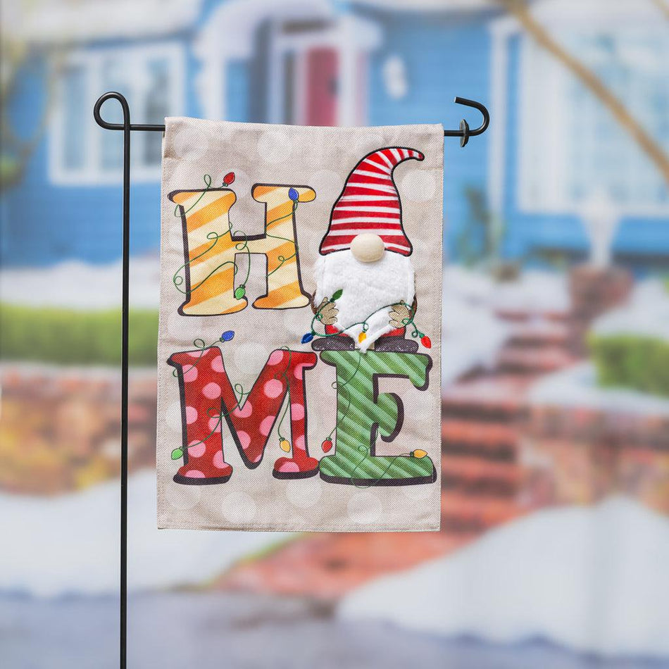 The Home Gnome garden flag features the word "HOME" in holiday colors with a gnome replacing the "O" and holding a string of lights. 