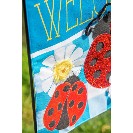 The Ladybug Plaid Welcome house banner features a trio of ladybugs on a bright blue checked background with white daisies and the word "Welcome". 