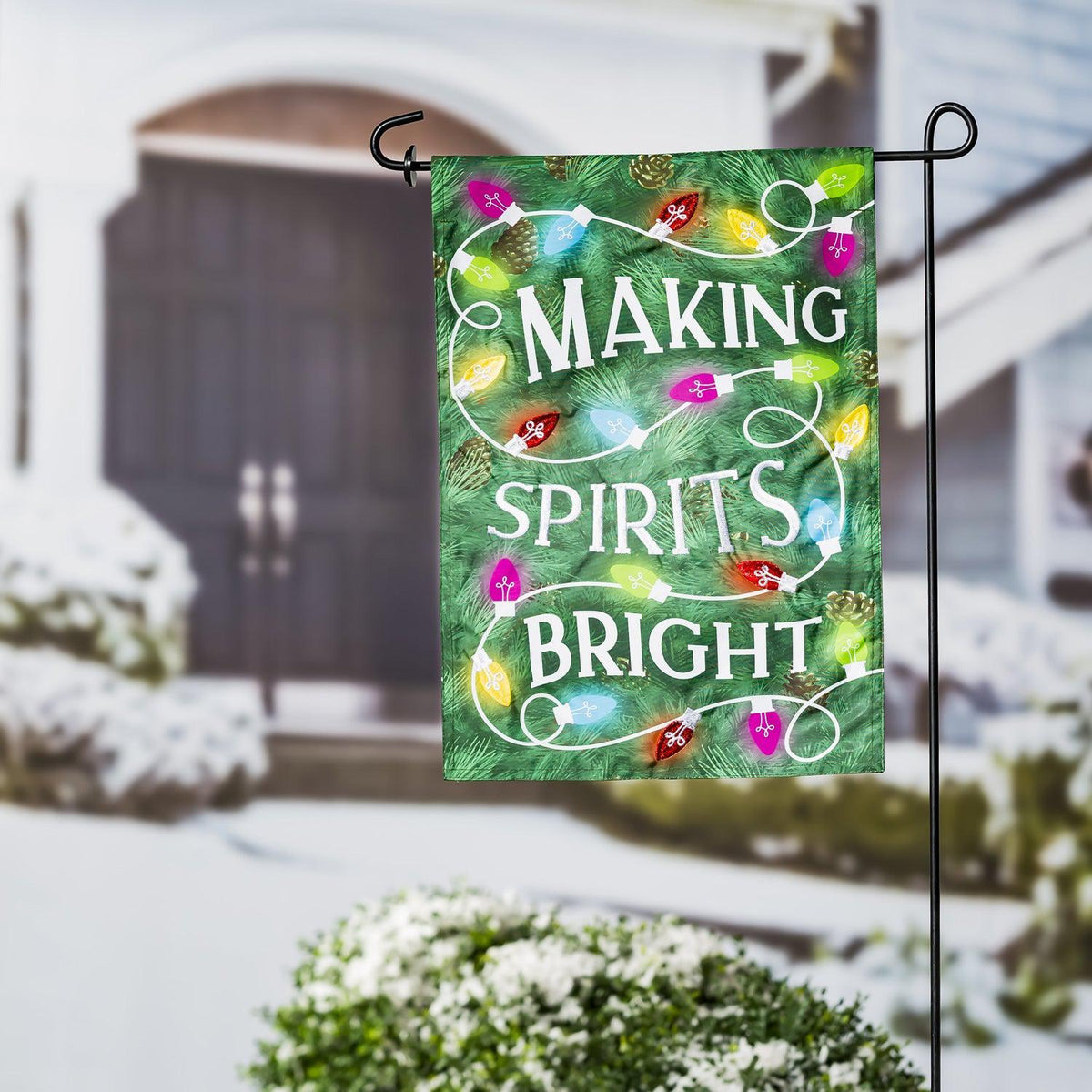 The Making Spirits Bright garden flag features a green pine needle background with a string of brightly colored Christmas lights.