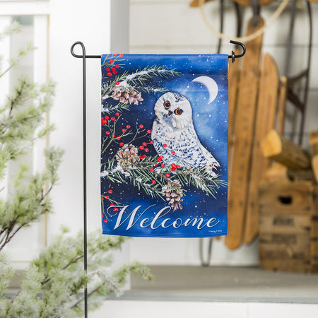 The Owl's Winter Greeting garden flag features a snowy owl at night resting on a pine branch along with the word "Welcome". 