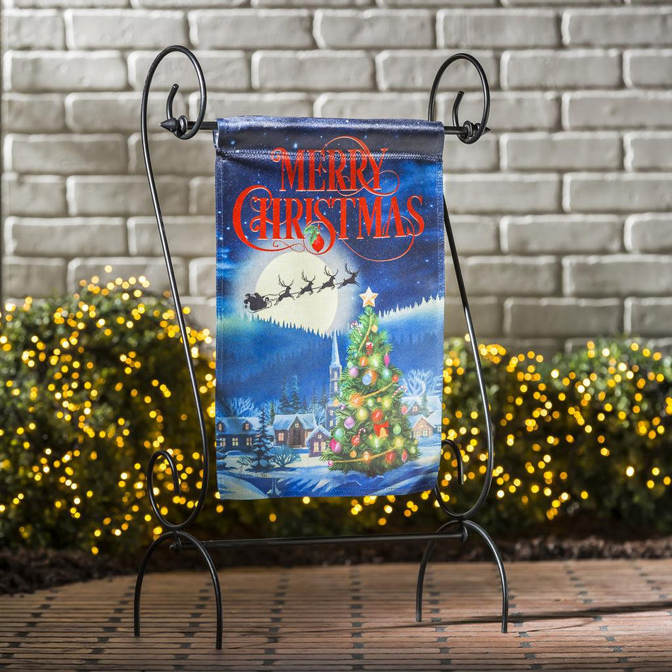 The Santa's Sleigh on Christmas garden flag features Santa and his reindeer flying over a small village with a beautiful Christmas tree, and the words "Merry Christmas" across the top. 