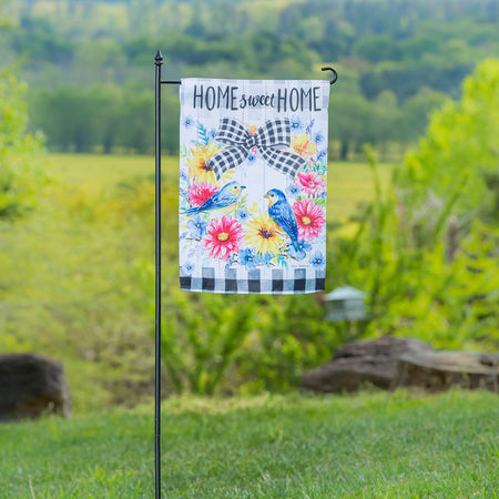 The Spring Bird Wreath garden flag features a spring floral wreath with a pair of bluebirds, black checked accents, and the words "Home Sweet Home".